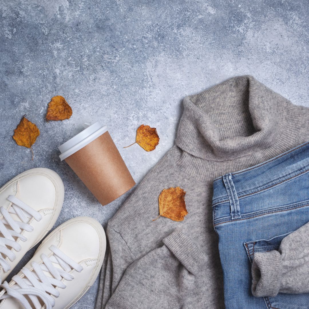 image of coffee cup, blue jeans, a turtle neck sweater and white sneakers among fallen leaves to signify the fall season