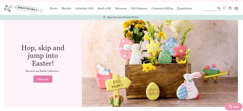 A screenshot of Biscuiteers’ website displaying the menu on top and a pastel-colored Easter-themed banner promoting their sale with the CTA “Hop, skip and jump into Easter” on a pale pink background on the left side and a wooden box filled with Easter-themed cookies on the right