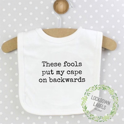 White cotton baby bib that reads “These fools put my cape on backwards”