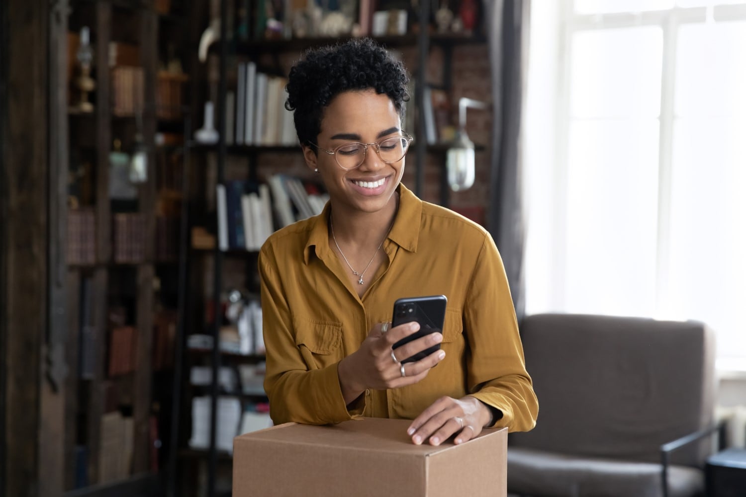 A women checks her smartphone while holding a boxed package in her home.