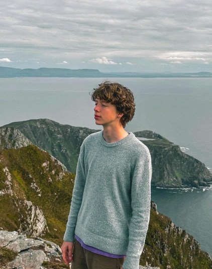 a man in a blue sweater standing in front of a cliff and body of water