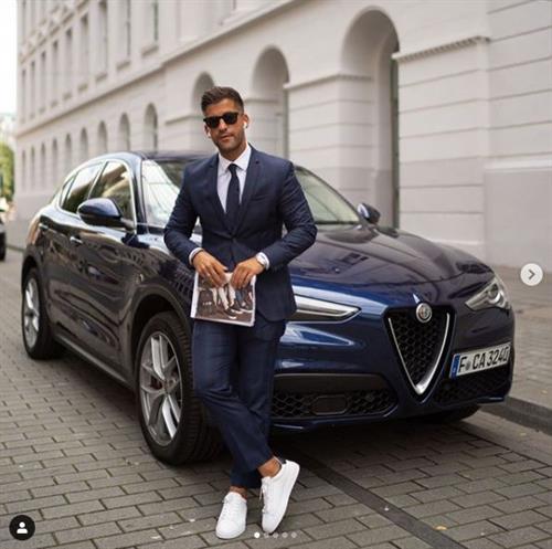 German influencer Kosta Williams wearing a suit and sneakers and leaning against a sports car
