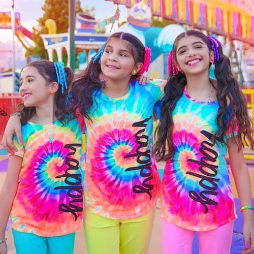The Alj Sisters wearing rainbow tie dye shirts with the words “Happy” in black letters in front of a amusement park