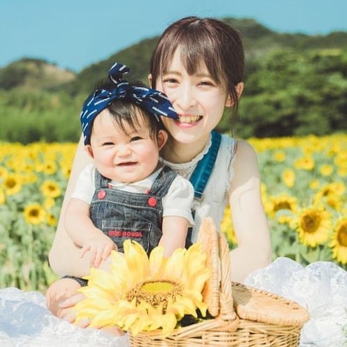 Satomi and Aichi wearing white clothes and overalls in front of a field of sunflowers and mountains