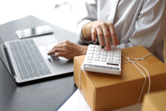 Person calculating international shipping costs using a calculator and a laptop computer.