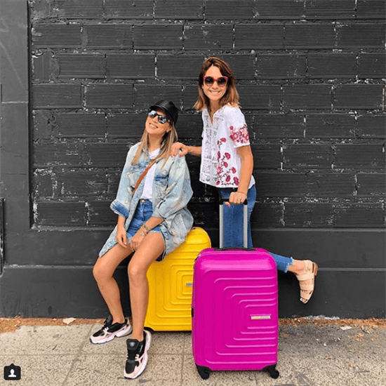 Bloggeres Coty Crotto and Maru Gandara standing next to black wall holding pink and yellow suitcases