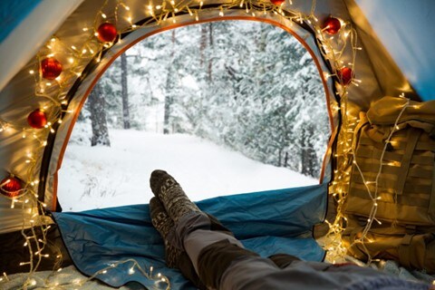 View from inside a camping tent. The entrance, lined with ornaments and lights, is zipped open revealing a snowy forest. A person inside the tent reclines with their sock-covered feet pointing towards the forest outside the tent.