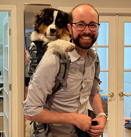 a man in glasses smiling with a black and white dog in his backpack