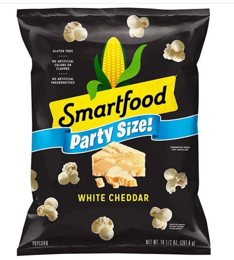 A black party size of Smartfood’s White Cheddar popcorn with the yellow Smartfood logo with a corn on top in the middle, the party size ribbon in light blue underneath, a piece of white cheddar cheese below the ribbon, and popcorn pieces scattered all over