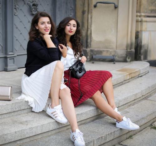 Sisters and influencers Samira and Laura sitting on outdoor steps in pleated skirts