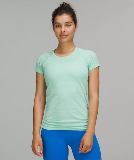 A woman with a Wild Mind Swiftly Tech Short Sleeve 2.0 Shirt and blue workout shorts in front of a grey background