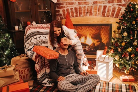  Two people in pajamas enjoy gifts, with a roaring fireplace and Christmas trees in the background