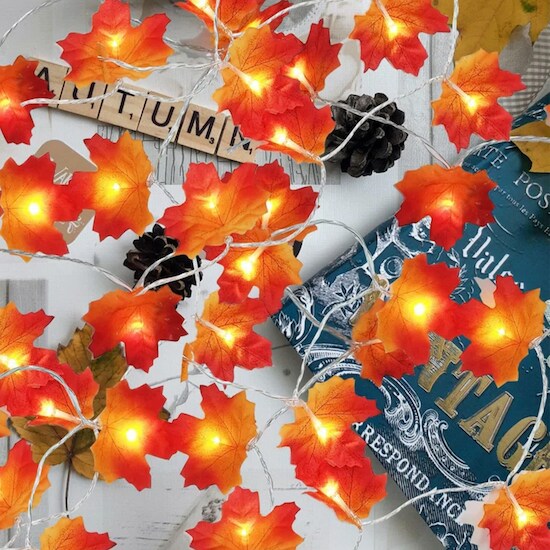 A set of orange Holiday Aisle String Lights in the form of leaves attached to a bendy wire on top of a table full of papers, covers, and scrabble tiles that spell the word “Autumn”