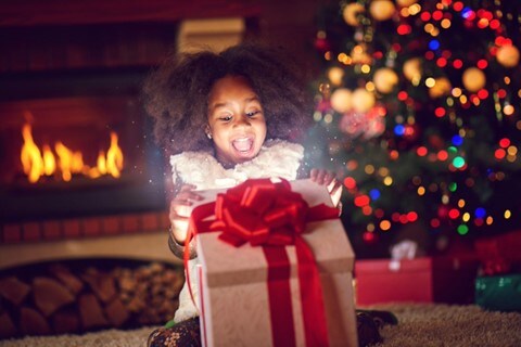 A little girl cracks open the lid of a present, light comes from the box, illuminating her face in front of a glowing fireplace and lit Christmas tree.