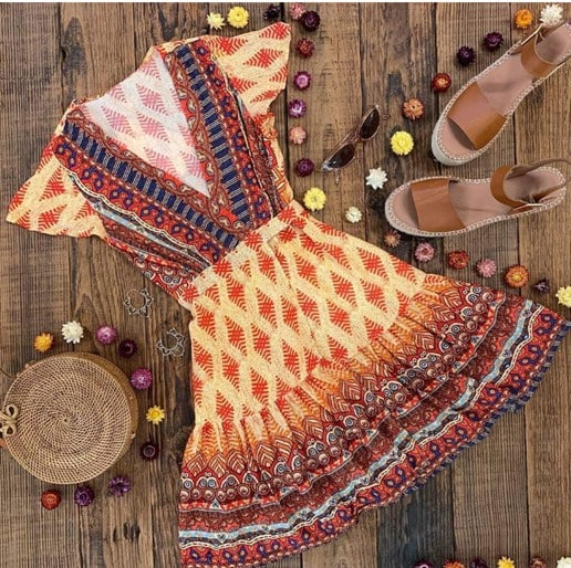 ZESICA’s bohemian-inspired dress in orange, mixed with red, yellow, and blue floral and baklava designs, laid on a wooden table. There are boho-inspired brown sandals and cat-eye sunglasses to the right, and a small round boho bag to the left side of the dress. Flowers surround each piece.