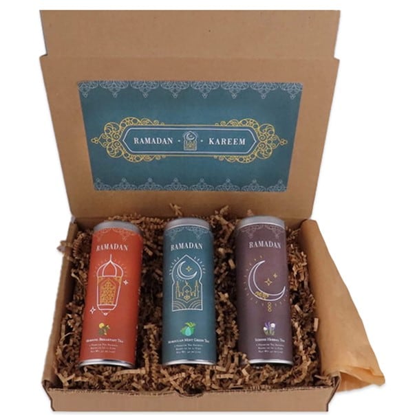 A square cardboard box filled in with packing materials and three different cans of tea – red, blue, and purple, all with Islamic motifs on them. There is a white, blue and gold print on the inside of the box that reads “Ramadan Kareem”.