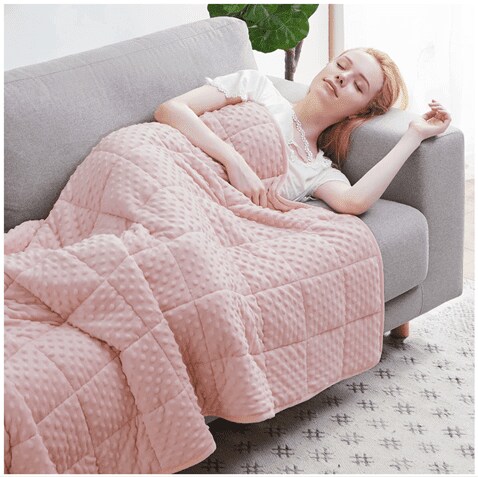 A blond woman sleeping on a gray couch, covered with a pink weighted blanket from Huloo