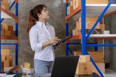 	A woman holding a tablet surveys shipping packages on shelves around her.