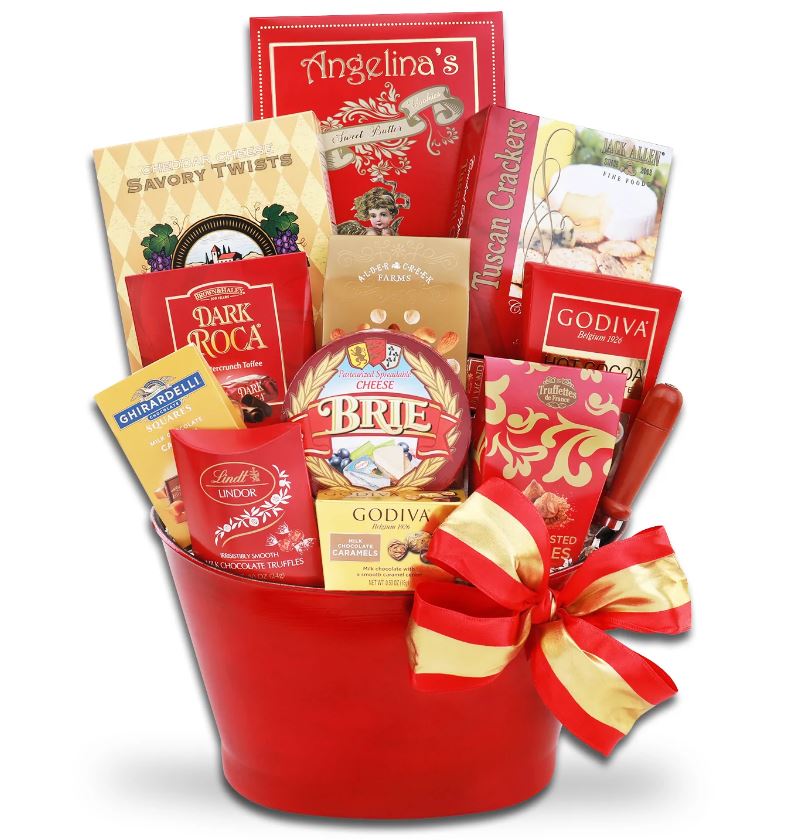 Red gift basket with a red and gold bow with an assortment of cookies, cheese, crackers, and sweets. 
