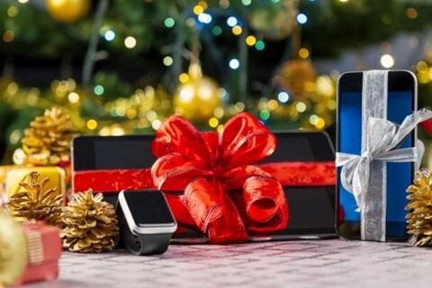 Close up on a smartphone, smart watch, and tablet, sitting in front of a decorated Christmas tree. Each device is wrapped in ribbon with a large bow.