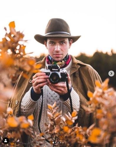 Icelandic influencer and photographer Joe posing in a field with his camera in a brown fedora and jacket 