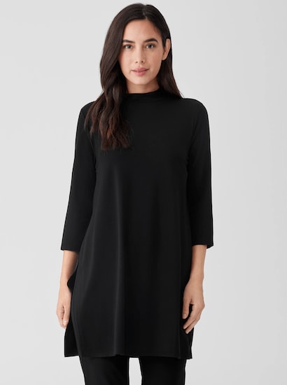 A woman wearing a black Eileen Fisher Stretch Jersey Knit Mock Neck Long Top with slim black pants