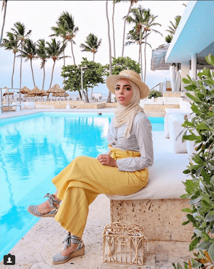 Blogger Heba Jalloul sitting next to pool wearing hat over white hijab and yellow pants