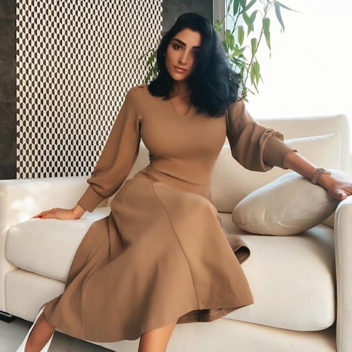 Daniella Debies wearing an elegant clay color long sleeve dress and a silver wrist watch on a white couch