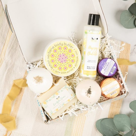 An open white self-care box for women containing artisan soap, bath bomb, bath salt, scrub, body butter, body wash, body lotion, and a shower steamer