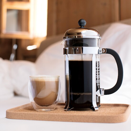 A stainless steel 3-Cup Bodum Chambord French Press and a cup of coffee on top of a beige tan board and a white blanket