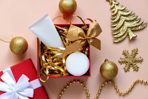 Open present box with gold Christmas ornaments scattered near the box. The box holds two cream beauty products cushioned by gold tinsel.
