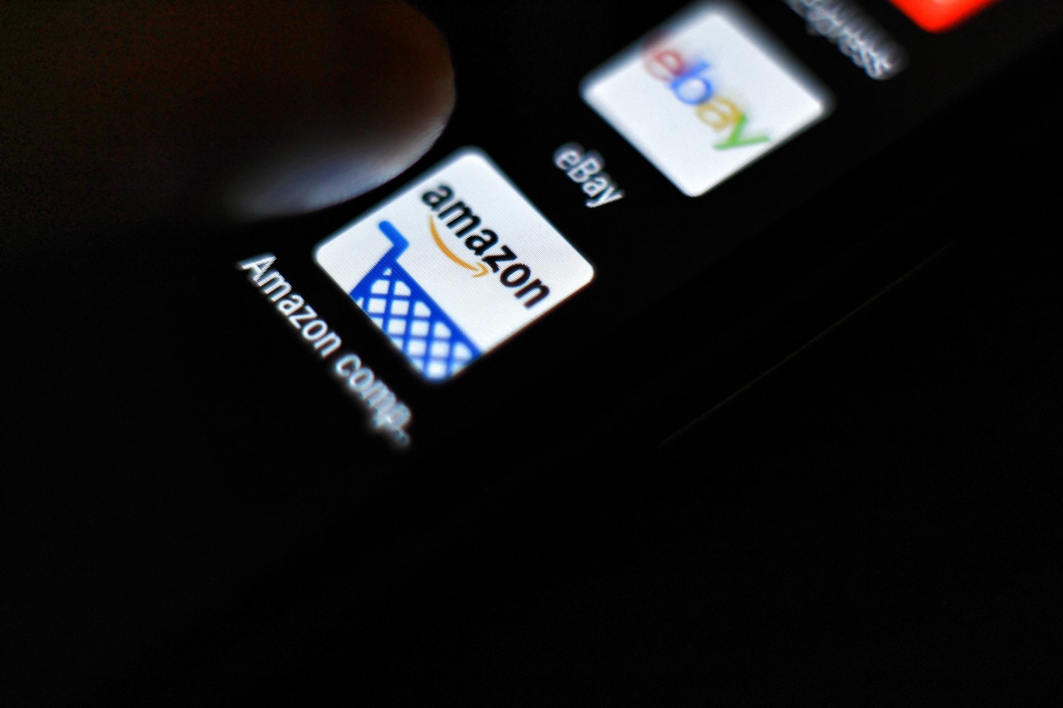 A dark smartphone screen displays the eBay app stacked above the Amazon app. A finger hovers next to the Amazon app