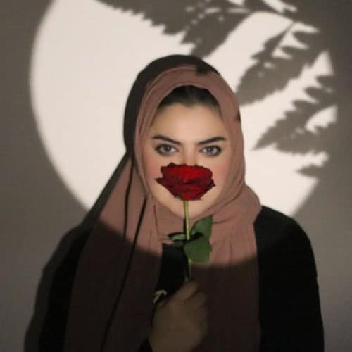 Jay Sajer wearing a brown hijab and black long sleeve shirt, holding a rose in front of a spotlight with a tree leaf shadow