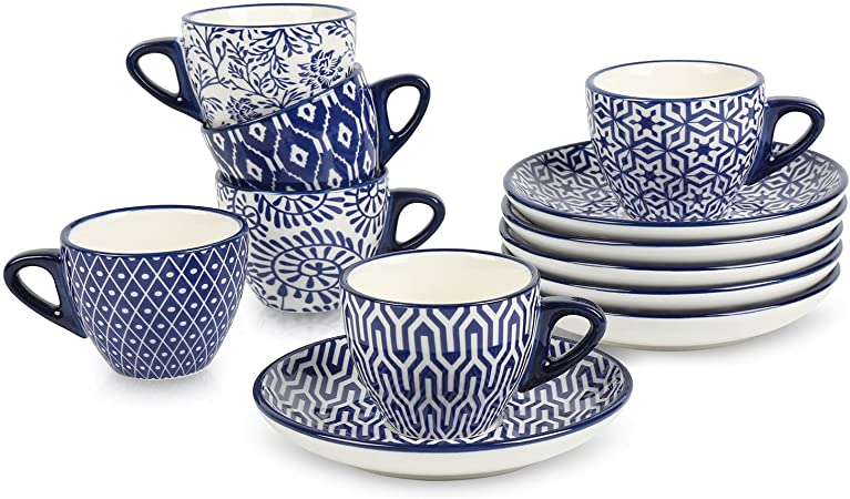 Six white cups and six saucers with blue patterns from the Selamica Ceramic 2.8 oz Espresso Cups and Saucer Set on a white table next to a coffee pitcher 