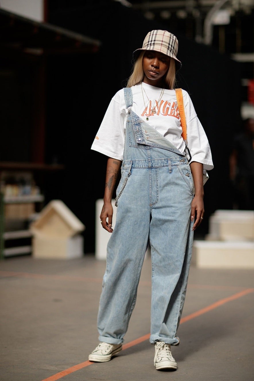 Model wearing a 90s-themed overalls with one shoulder off, a white, baggy t-shirt, white Converse, and a pleated bucket hat