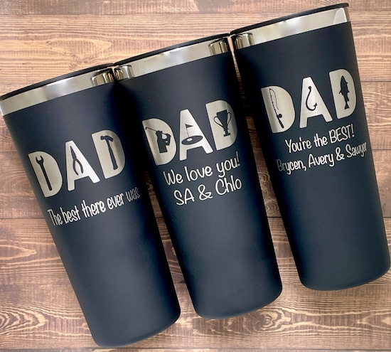 Three navy StitchedandLaseredCo Father’s Day Personalized Tumblers with white words and art print in front of a wooden panel